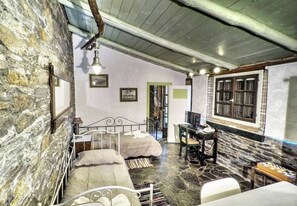 A rustic room with a stone wall, a bed with a metal frame, a desk with a chair, and a window with wooden shutters. - Kypri Apartments (Andros)