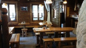 Uma cabana rústica de madeira com várias mesas e bancos, grandes janelas com cortinas e um interior acolhedor.