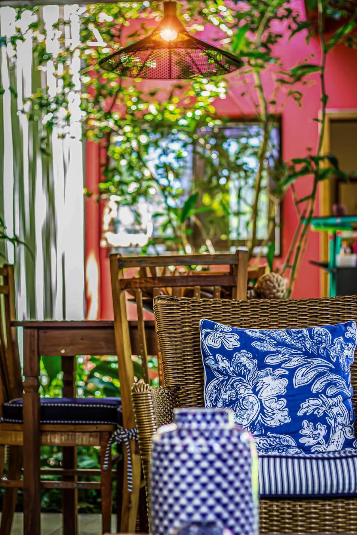 Uma cadeira de vime com almofada estampada em azul, um candeeiro pendente e uma estante com livros e objetos decorativos.