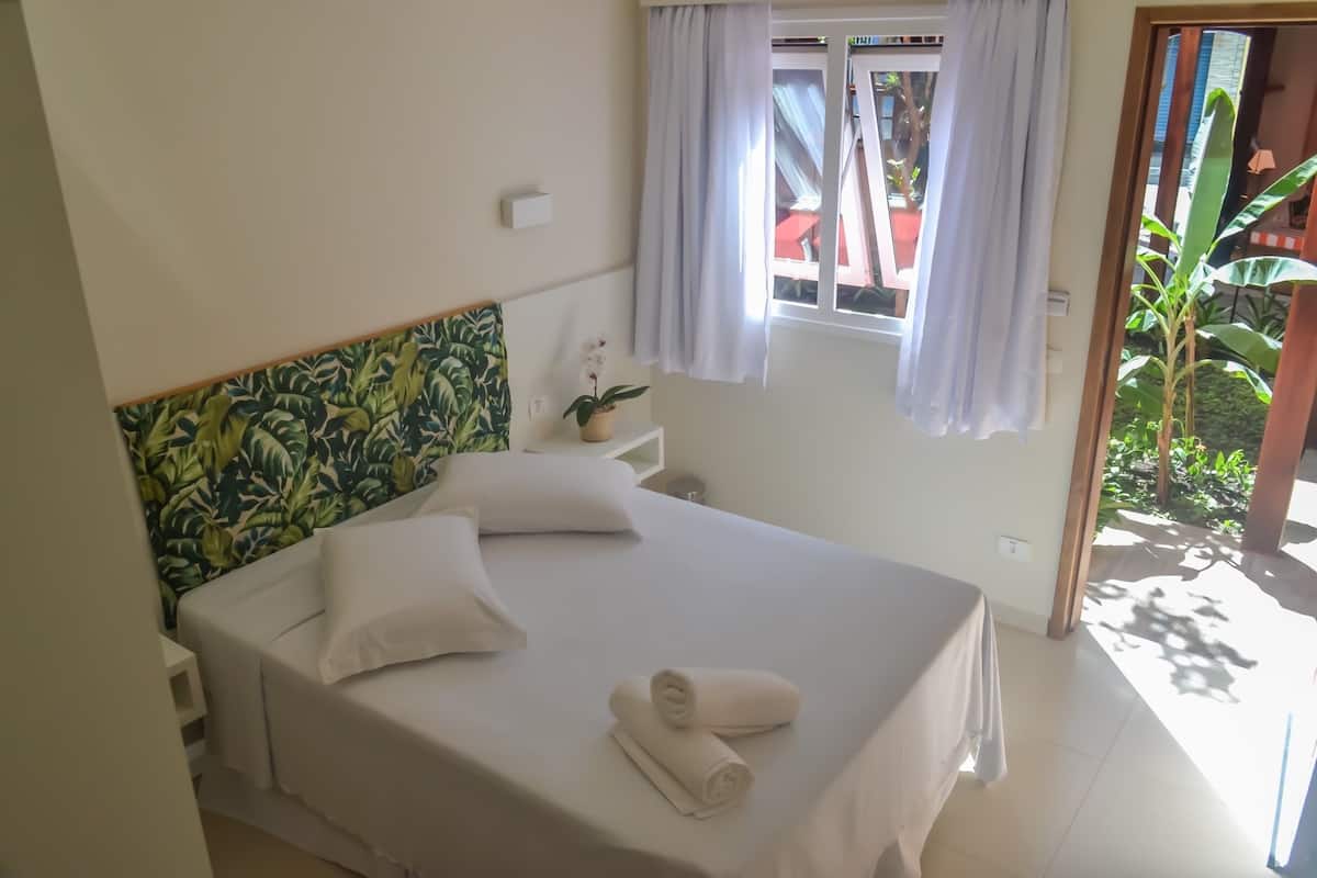 A neatly made bed with white linens and rolled towels, a small bedside table with a potted plant, and a window with curtains.
