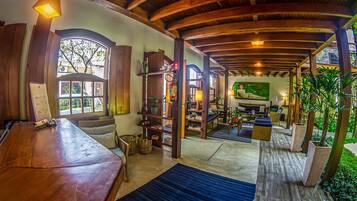 Una habitación amplia con techo de madera, una gran mesa de madera, un sofá y una estantería. Se ve una ventana con vistas a la vegetación.