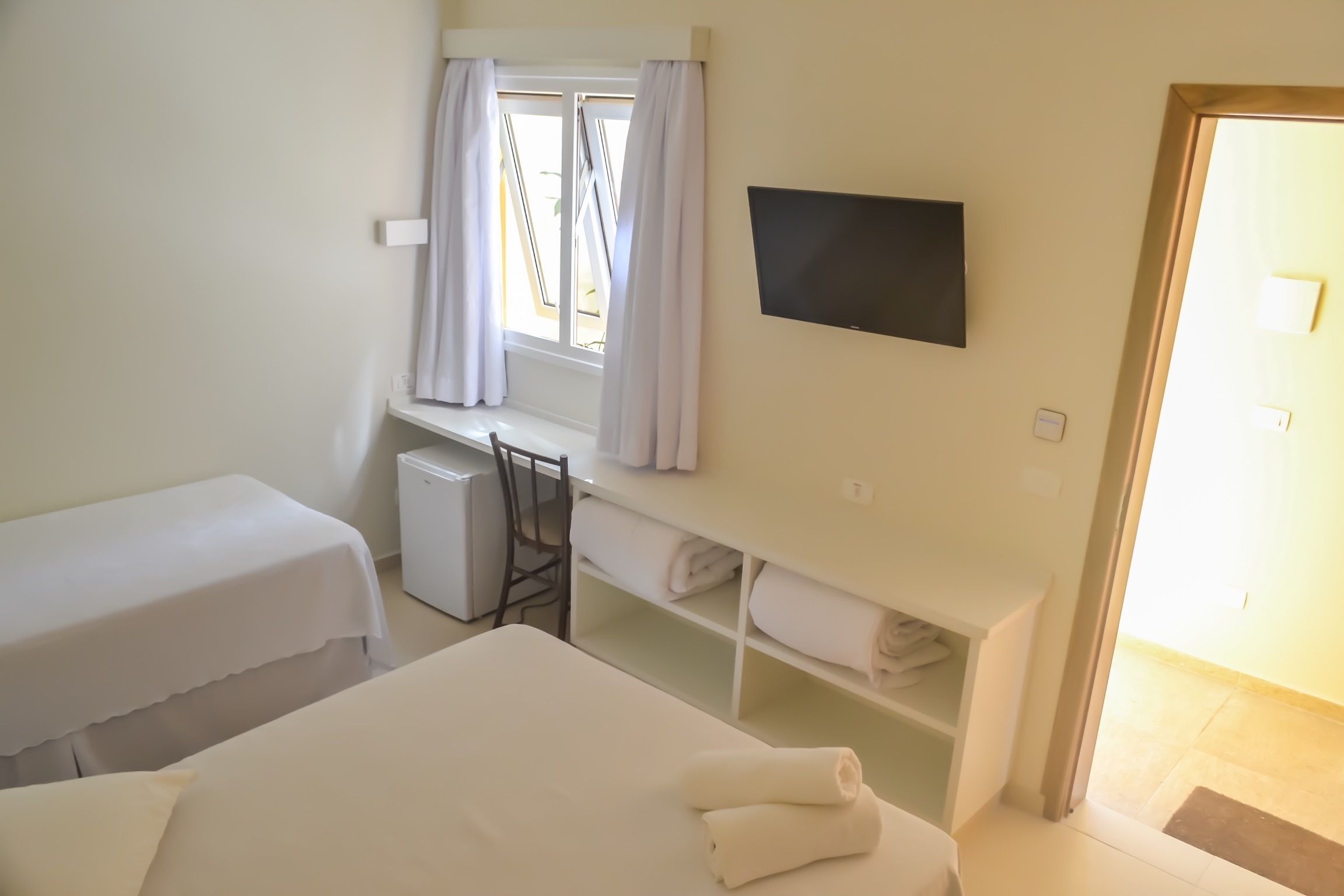 A hotel room with two beds, a desk with a chair, a TV mounted on the wall, and a window with curtains.