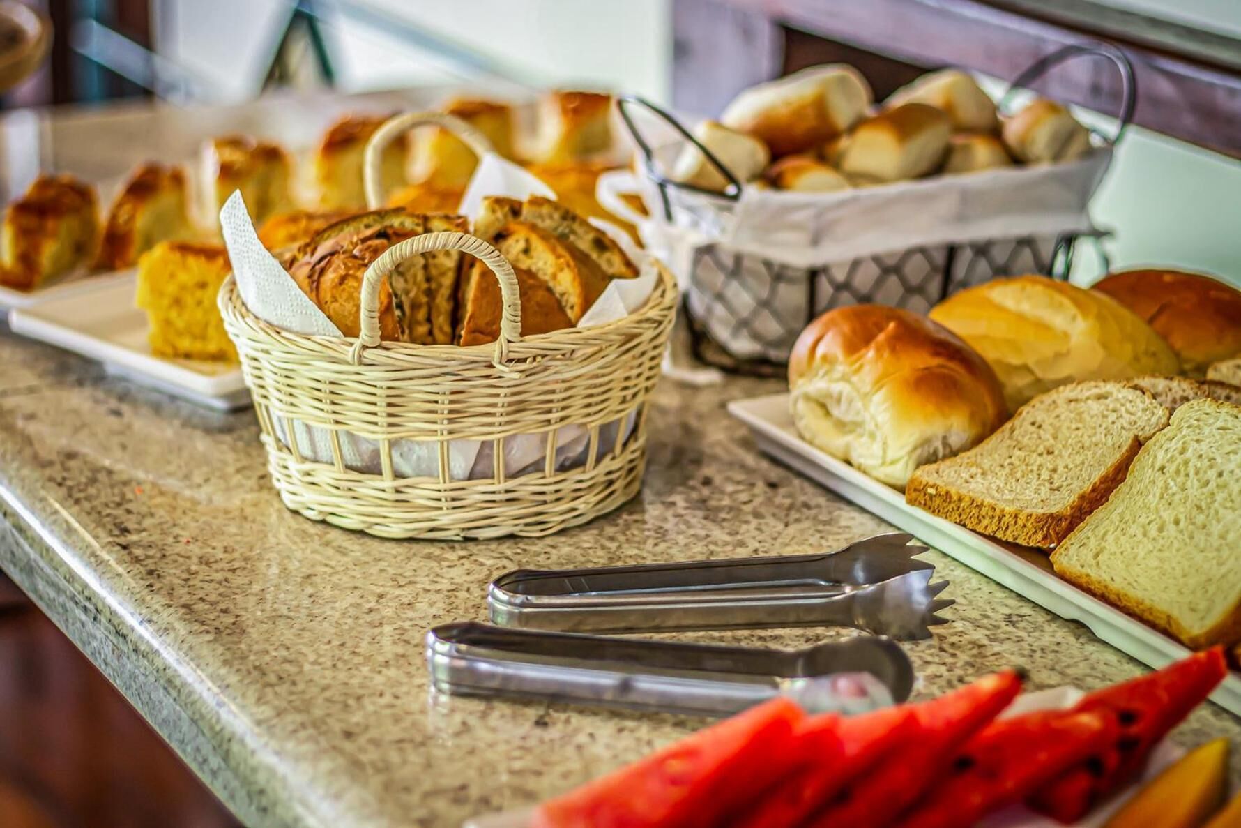 Uma mesa de buffet com diversos pães e pãezinhos, incluindo uma cesta de vime e uma bandeja de metal.