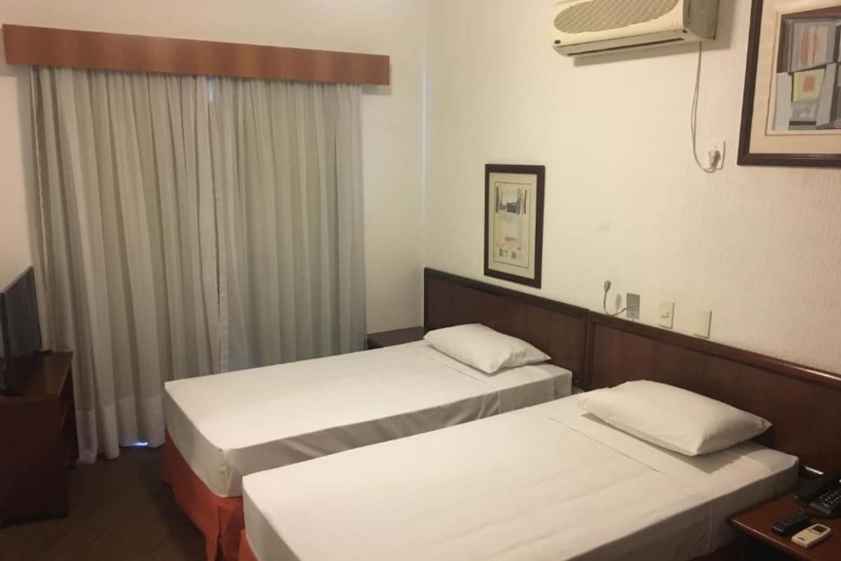 Ein Hotelzimmer mit zwei Einzelbetten, einem Holzkopfteil, einem Nachttisch mit Telefon, einem gerahmten Bild an der Wand und einer Klimaanlage.