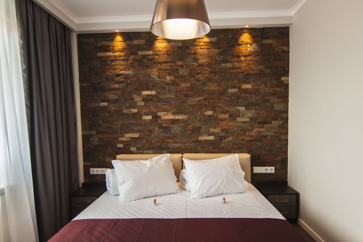 A neatly made bed with white linens and a red blanket, a wooden wall with a brick pattern, a hanging light fixture, and a dark nightstand with a power outlet.