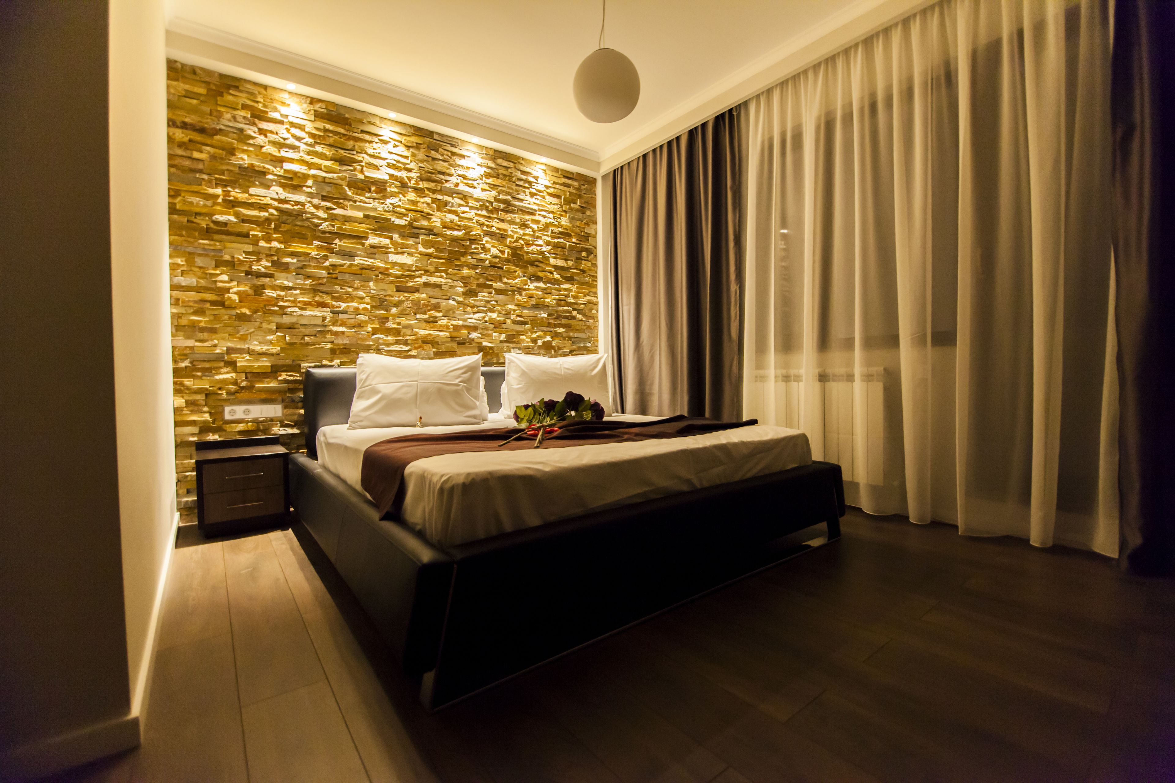 Un dormitorio moderno con pared de piedra, una cama grande, una mesita de noche y vistas al exterior a través de cortinas translúcidas.