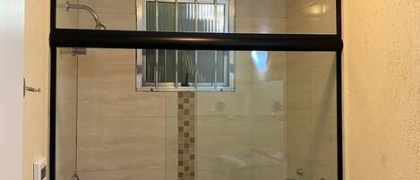 A glass-enclosed shower stall with a tiled wall, a small window, and a toilet visible to the right.