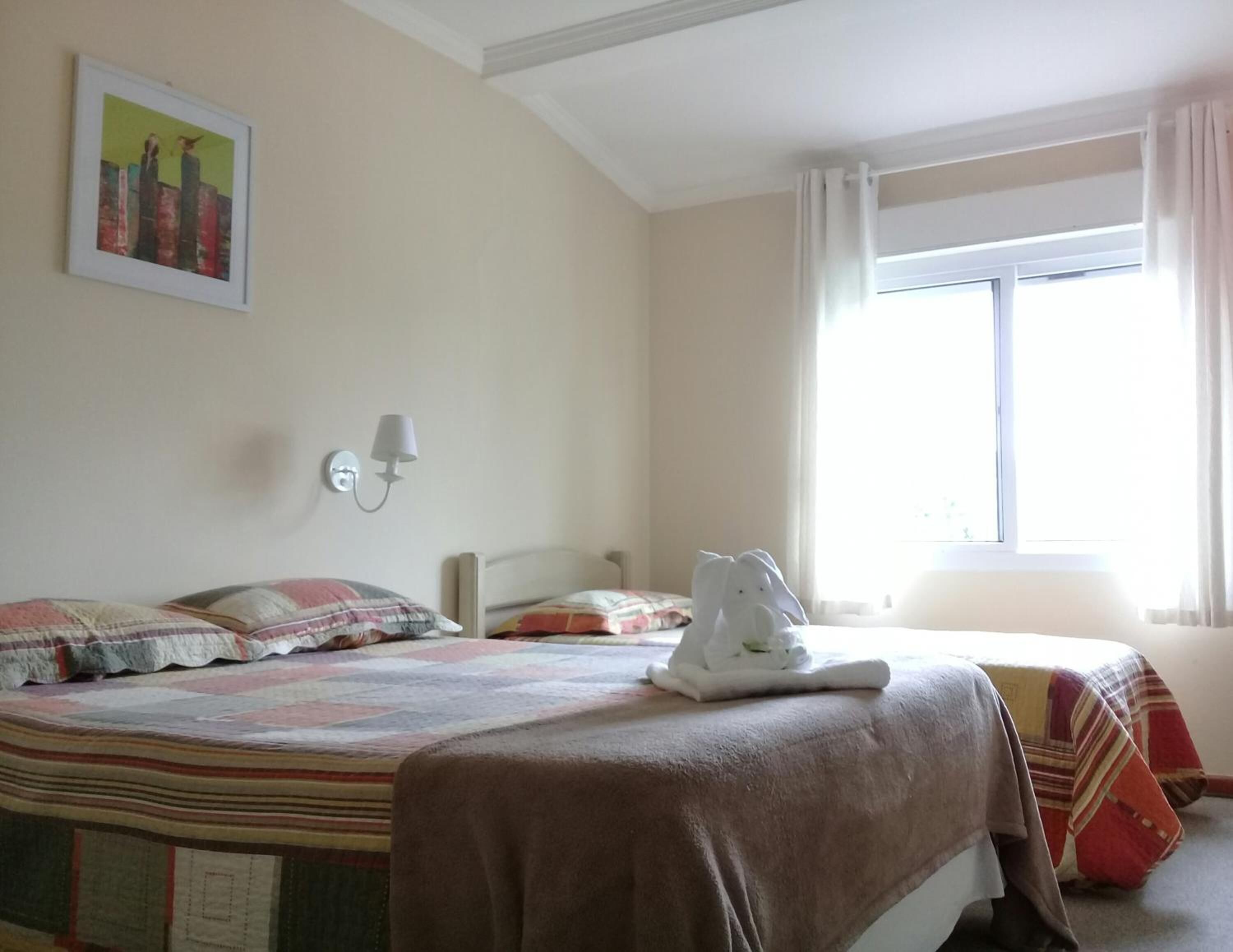 A hotel room with a bed, a nightstand, a wall-mounted lamp, a window with curtains, and a painting on the wall.