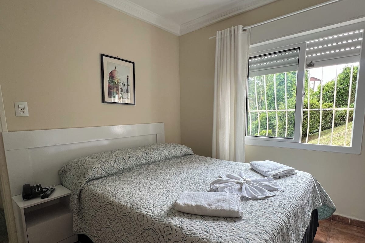Une chambre à coucher avec un lit, une table de chevet, une fenêtre avec des rideaux, un ventilateur de plafond et un tableau encadré accroché au mur.