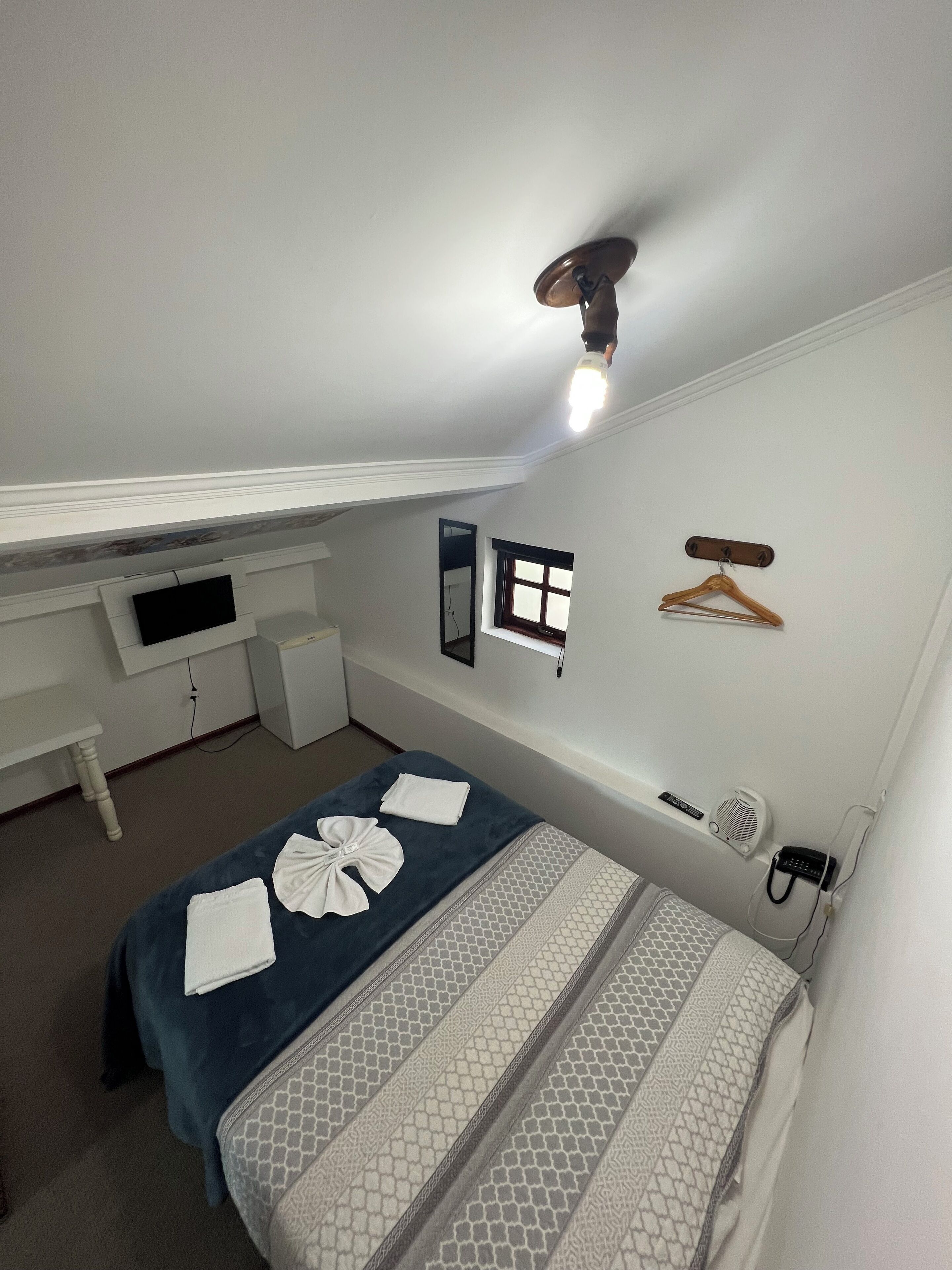 A bedroom with a bed, a desk, a chair, a hanging clothes rack, a small window, and a ceiling fan.