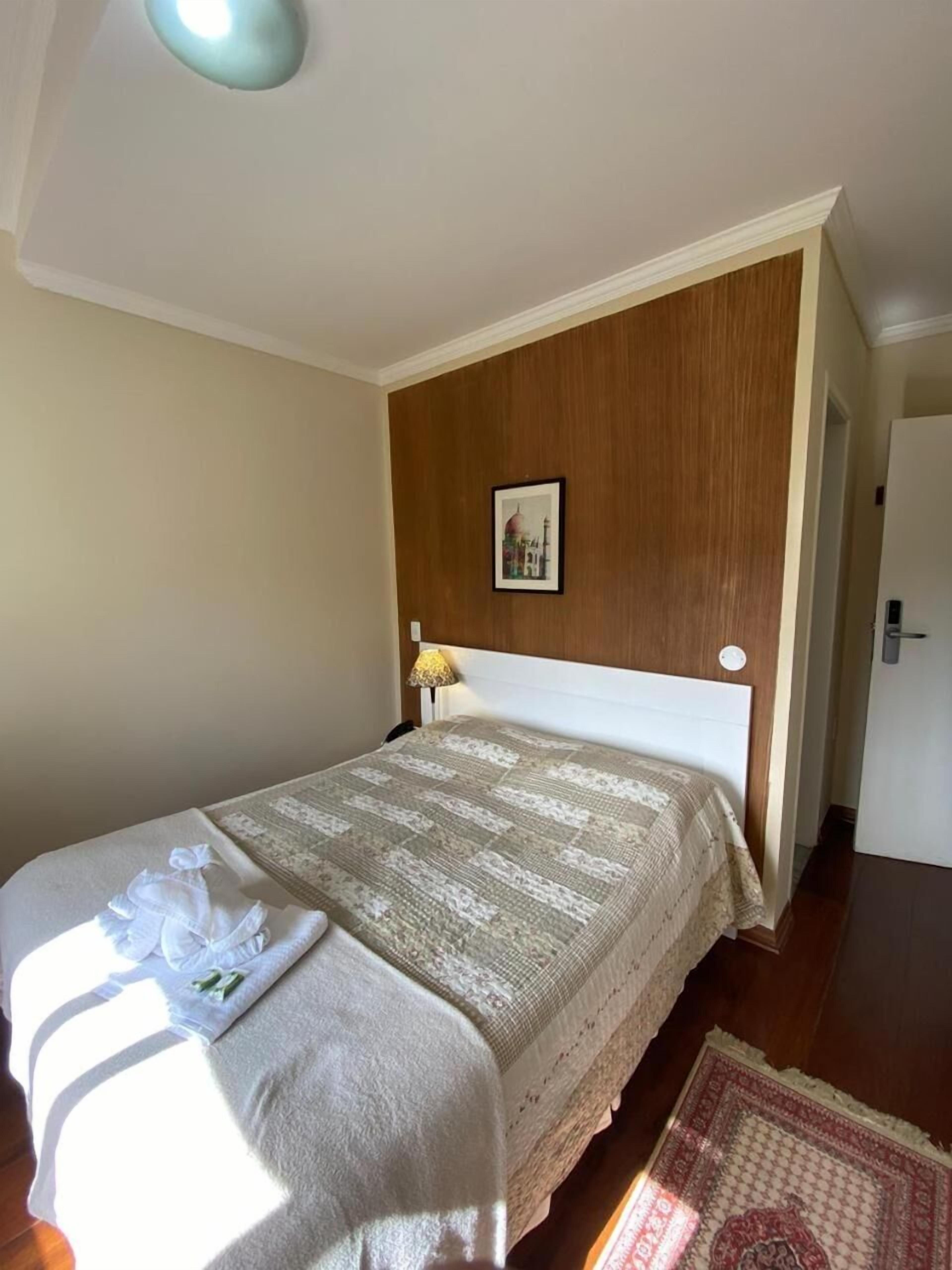 A neatly made bed with a patterned bedspread and a white pillow. A framed picture hangs on the wooden wall above the bed.