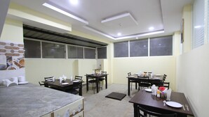 A dining area with tables and chairs, a kitchen area with a stove, and a window with blinds. - Tranzotel Bangalore Airport Hotel (Bengaluru)