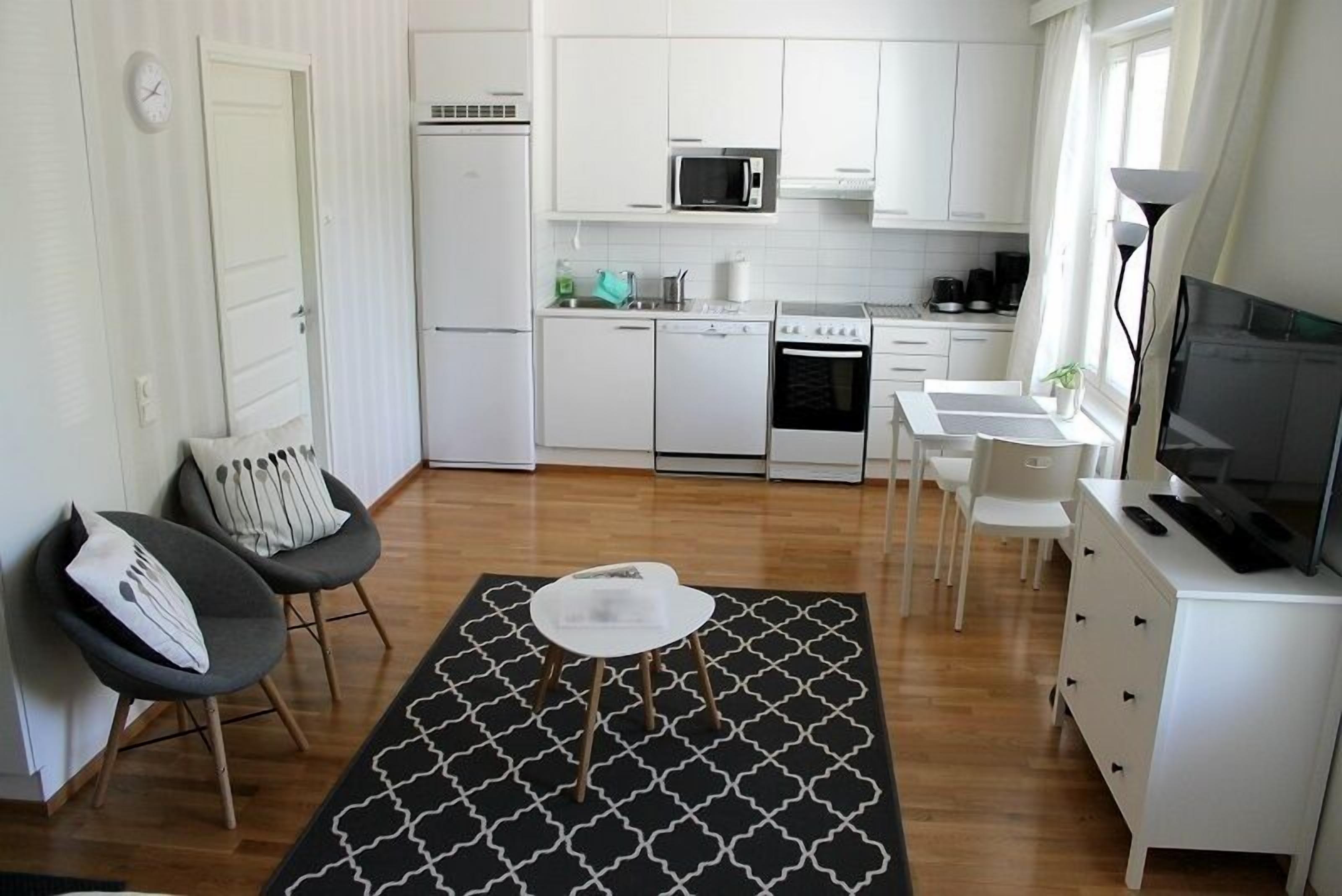 Une cuisine compacte avec des meubles blancs, un four à micro-ondes, une cuisinière et un lave-vaisselle. On y trouve un coin repas avec une petite table et des chaises, ainsi qu’un téléviseur posé sur un meuble blanc.