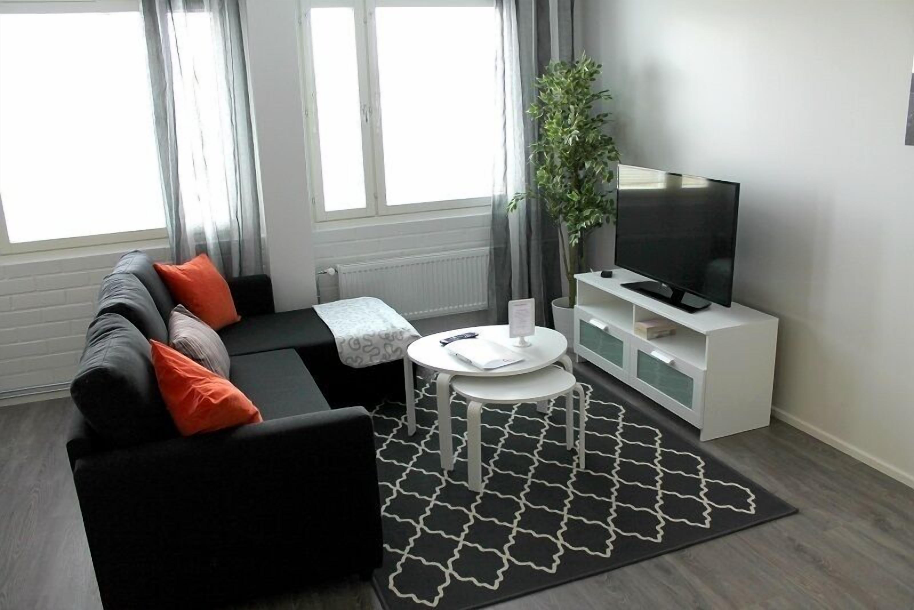 Una sala de estar moderna con un sofá negro, cojines naranjas, una mesa de centro blanca, un tapete con estampado blanco y negro, un mueble de televisión blanco y una planta en maceta.