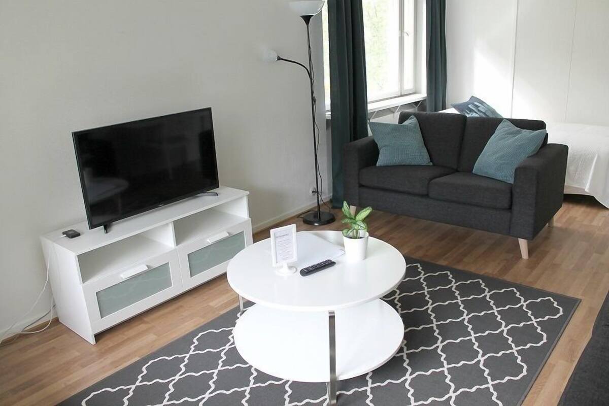 A modern living room with a flat-screen TV on a white stand, a grey sofa with teal cushions, a white coffee table, and a patterned rug.