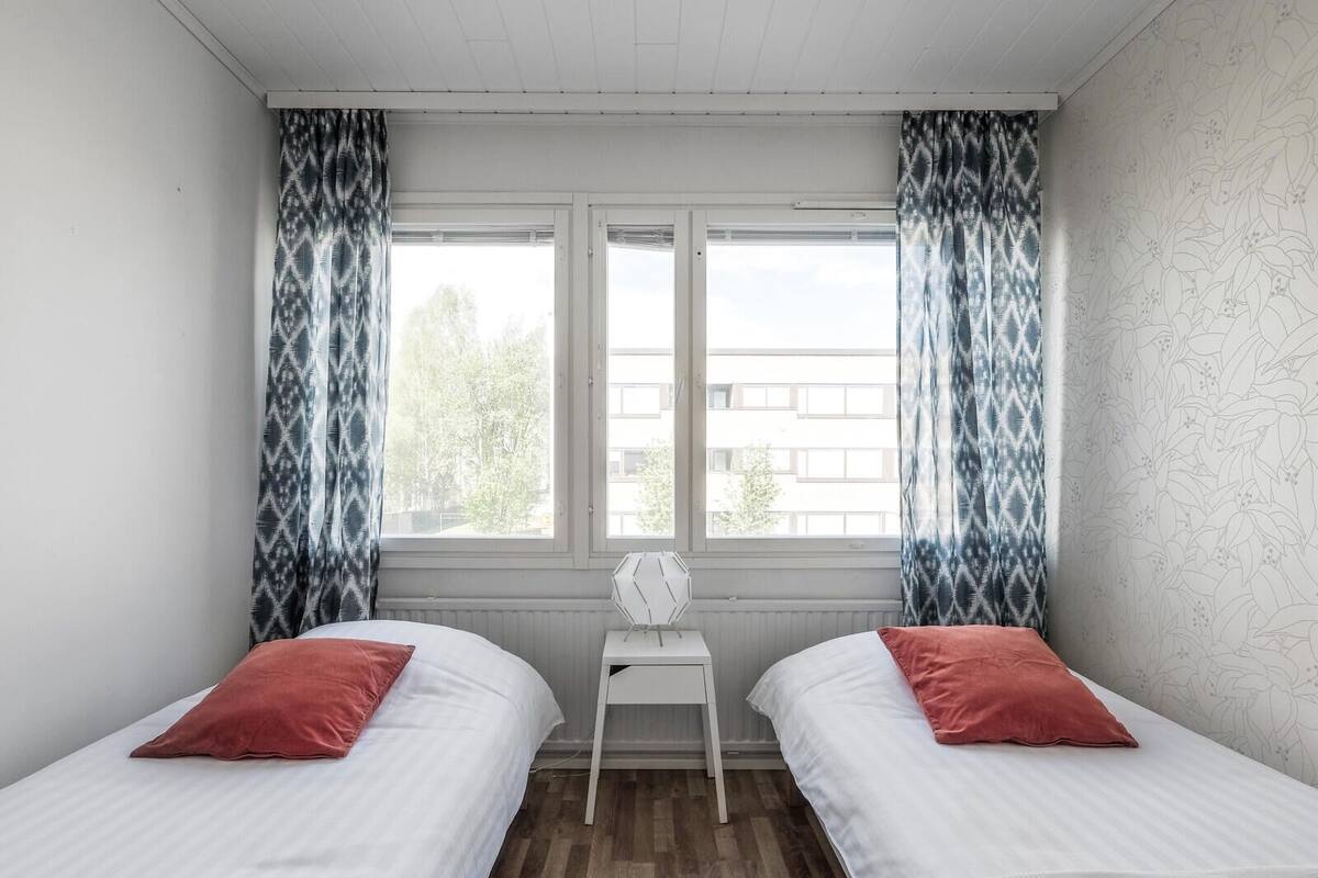 A room with two beds, a small white table, and a window with patterned curtains.
