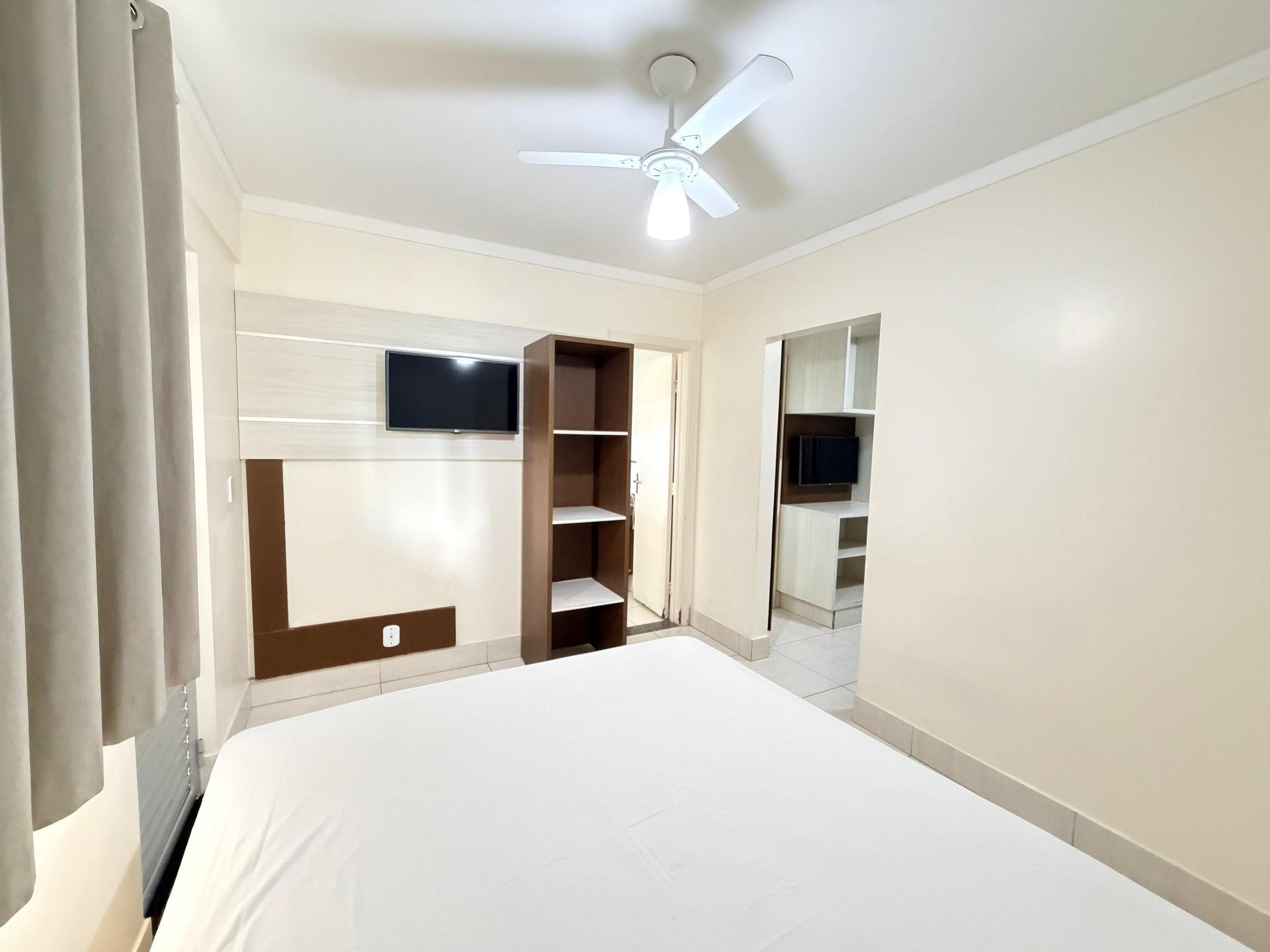 A bedroom with a bed, a TV mounted on the wall, a ceiling fan, and built-in shelving.