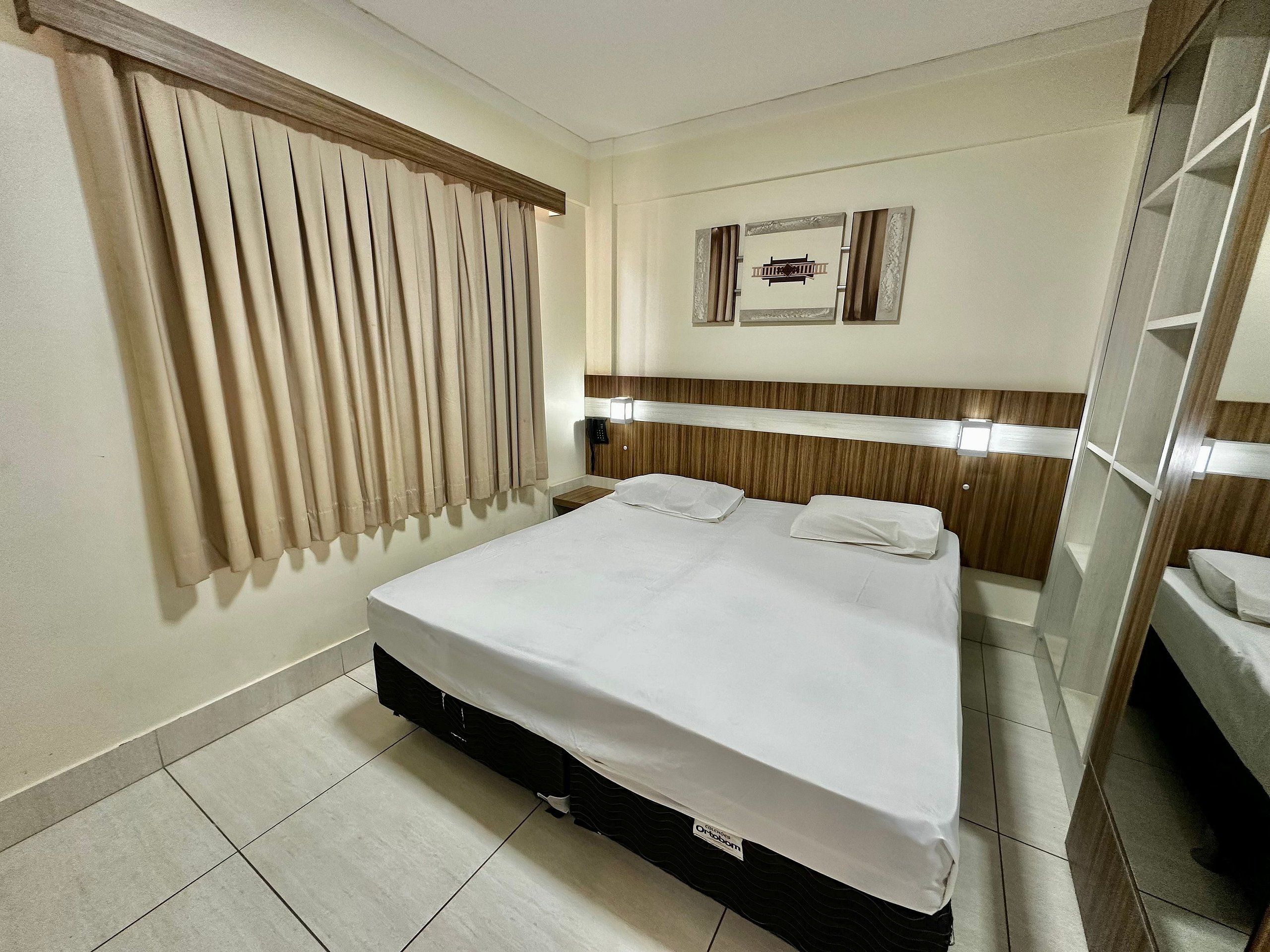 A hotel room with a bed, a nightstand, a mirror, a window with curtains, and a wall-mounted light fixture.