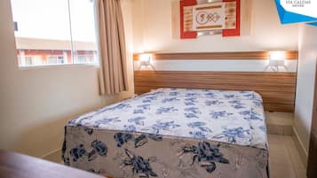 Hotellihuone, jossa on sänky, ikkuna verhoilla, seinälle ripustettu taideteos ja yöpöytälamppu.