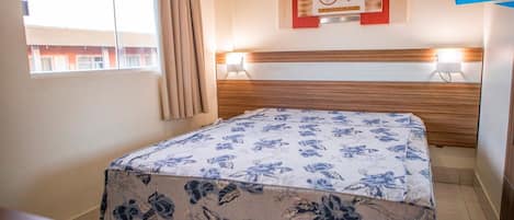 A hotel room with a bed, a window with curtains, a wall-mounted artwork, and a bedside lamp.