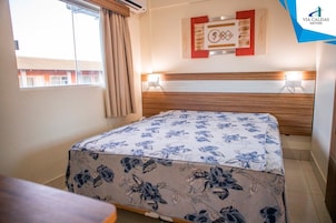Una habitación de hotel con cama, ventana con cortinas, una obra de arte colgada en la pared y una lámpara de noche.