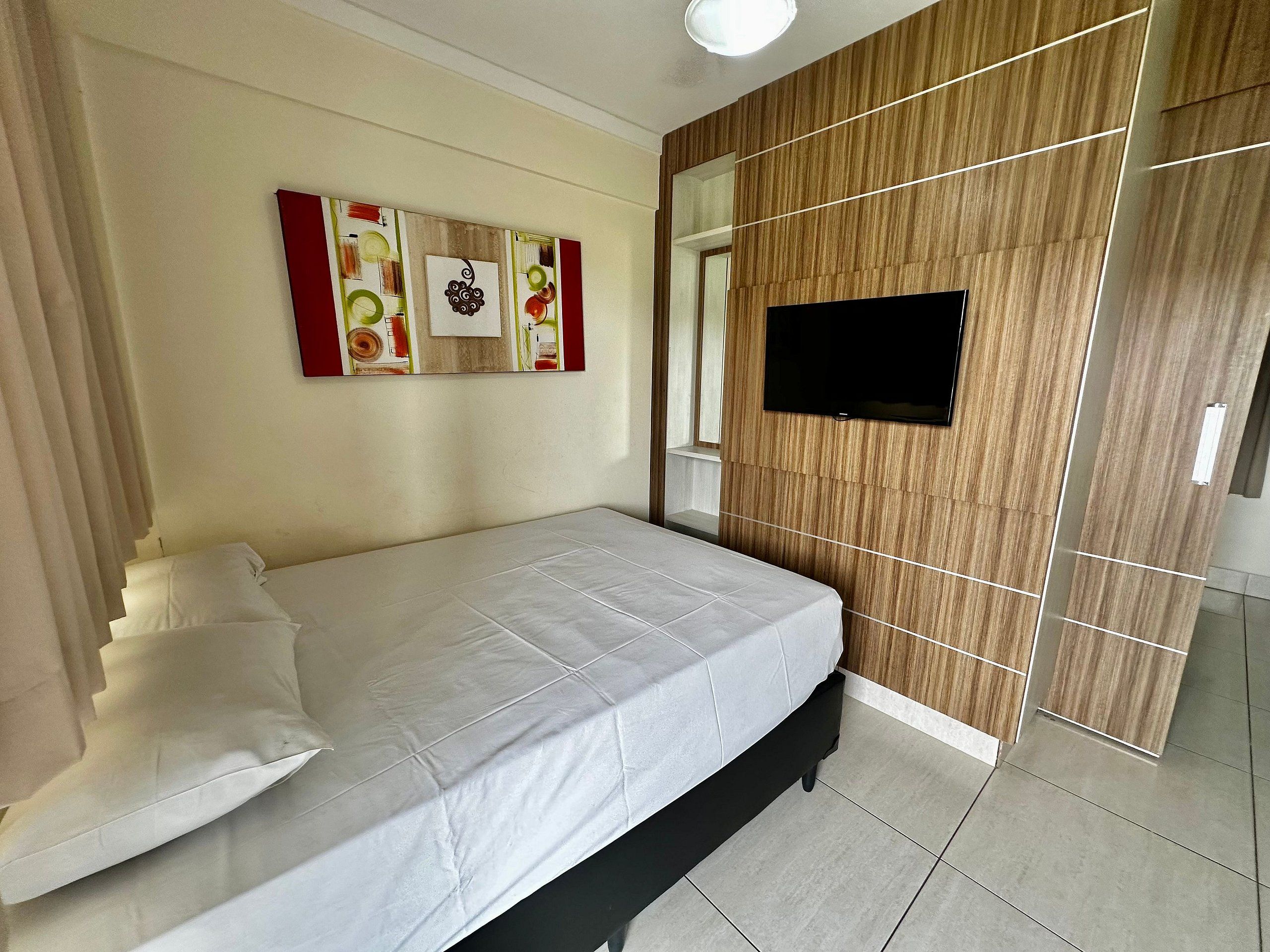 A hotel room with a bed, a flat-screen TV, a wall with a mirror, and a painting.