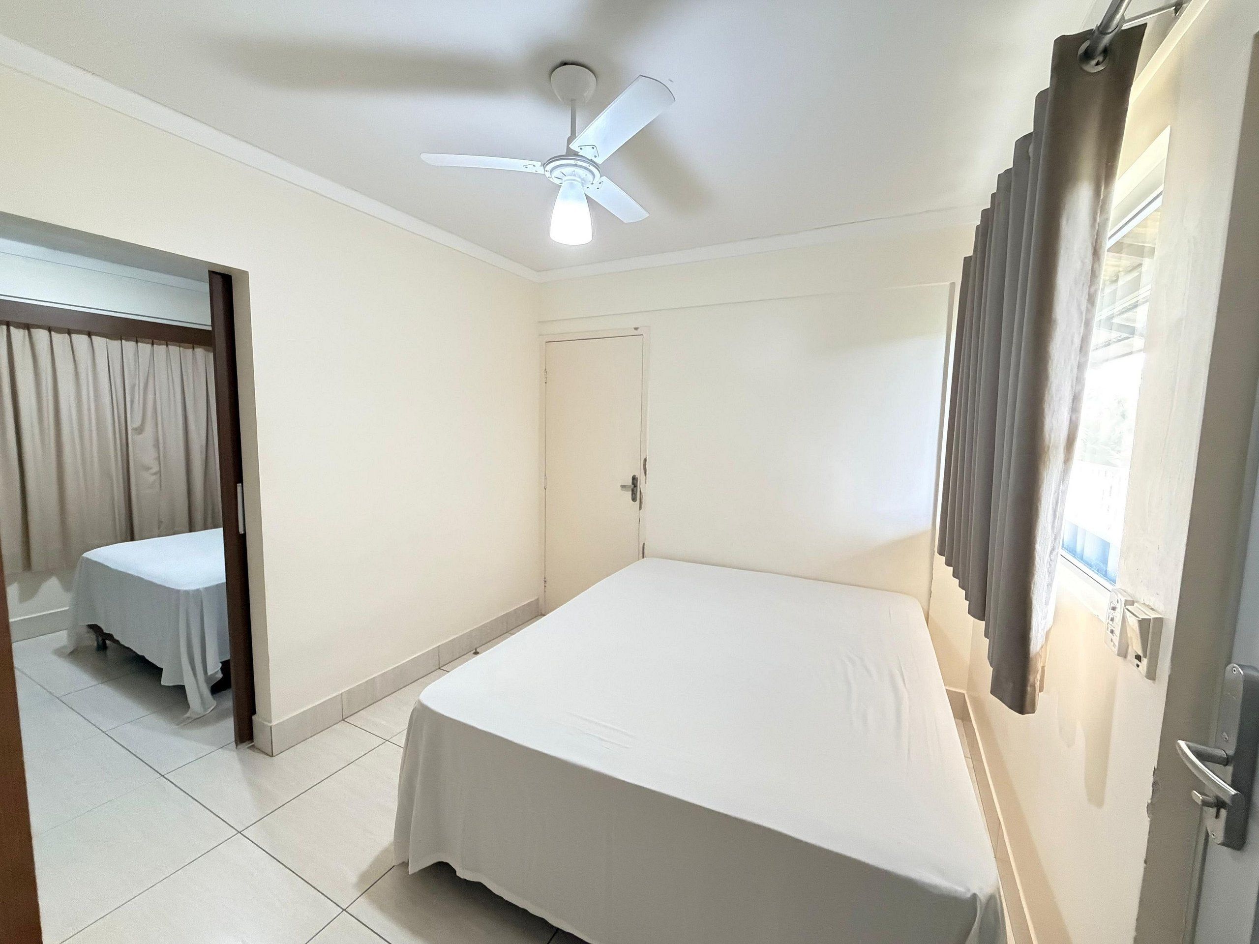 Quarto de hotel com uma cama de solteiro, ventilador de teto, uma porta e uma janela com cortinas.