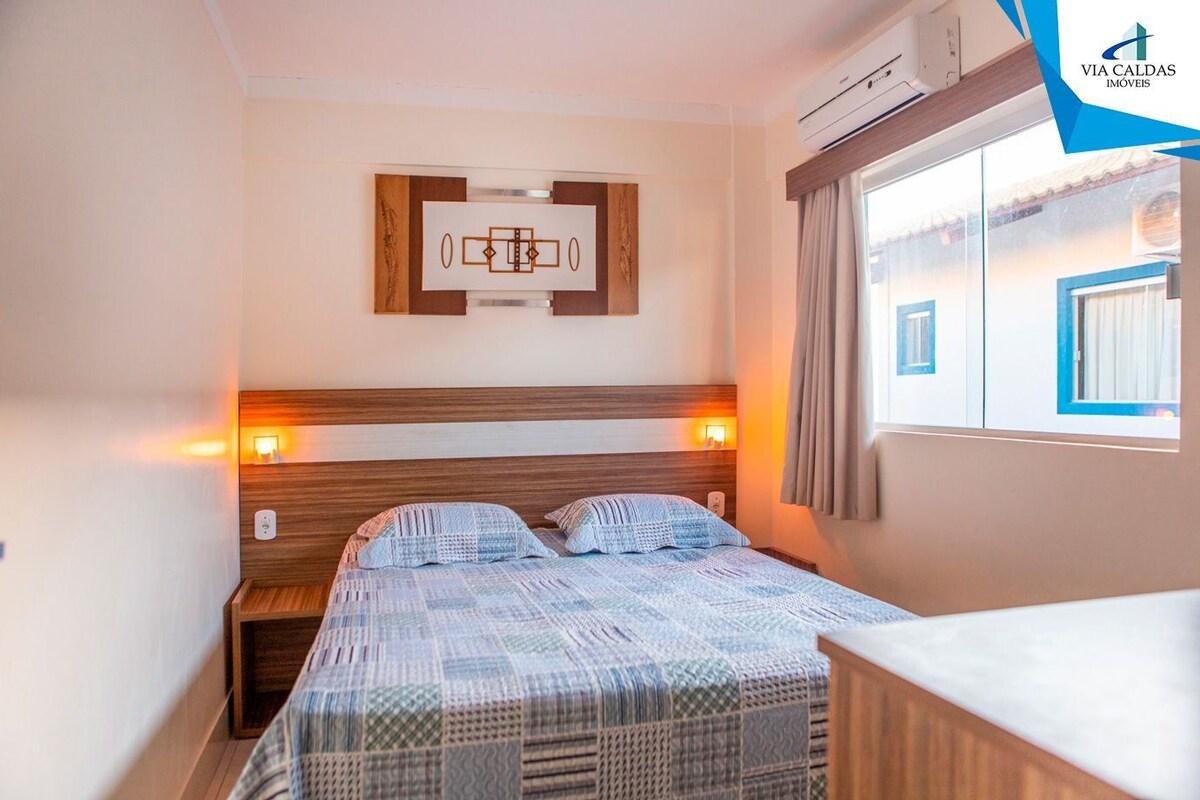 Una habitación de hotel con cama, mesitas de noche, un cuadro colgado en la pared, un equipo de aire acondicionado y una ventana con cortinas.