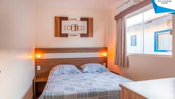 A hotel room with a bed, bedside tables, a wall-mounted artwork, an air conditioning unit, and a window with curtains.