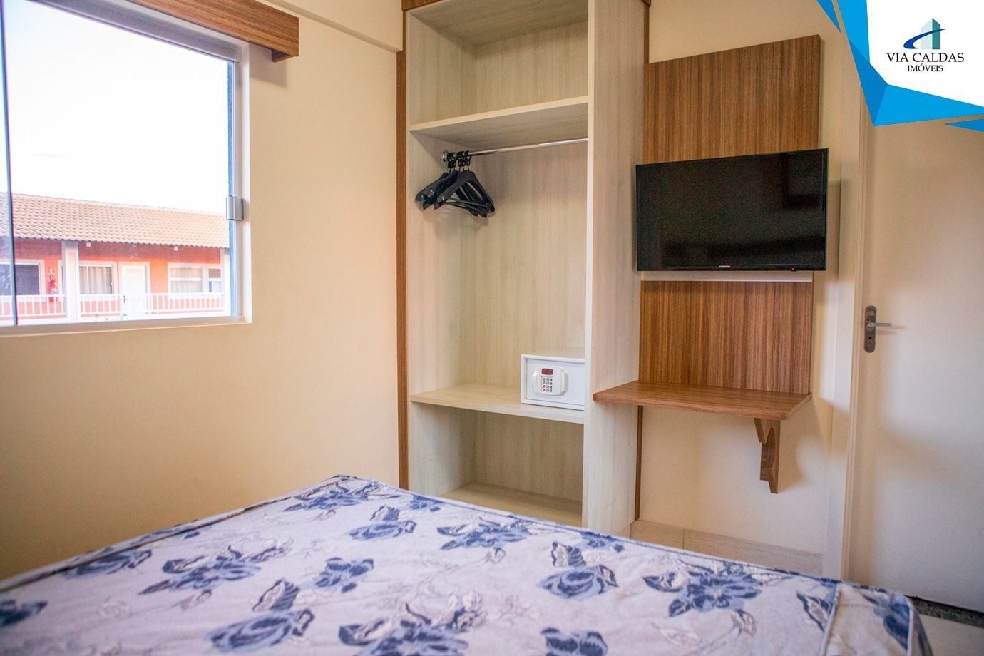 A bedroom with a bed, a TV mounted on the wall, a wardrobe, and a window with a view of buildings.