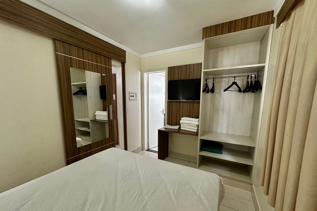 Una habitación de hotel con cama, armario con ropa colgada, televisor y espejo.