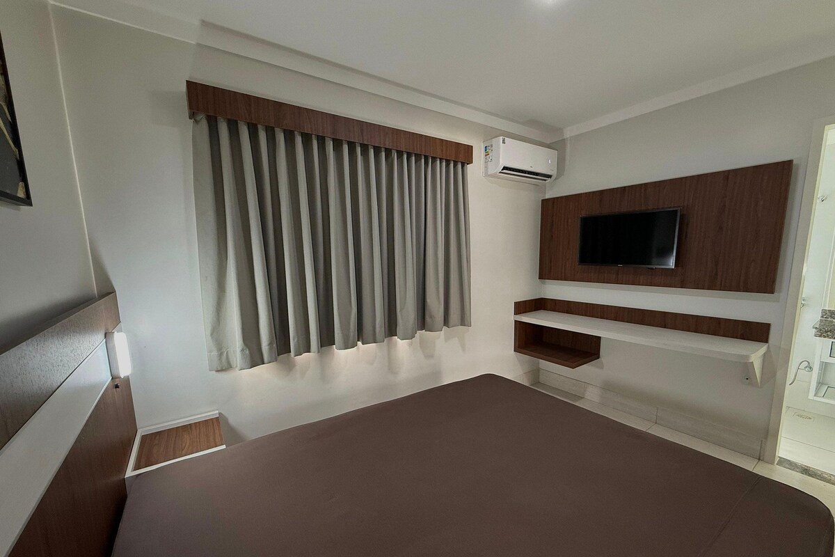 Un dormitorio con cama, televisor montado en la pared, ventana con cortinas y aire acondicionado.