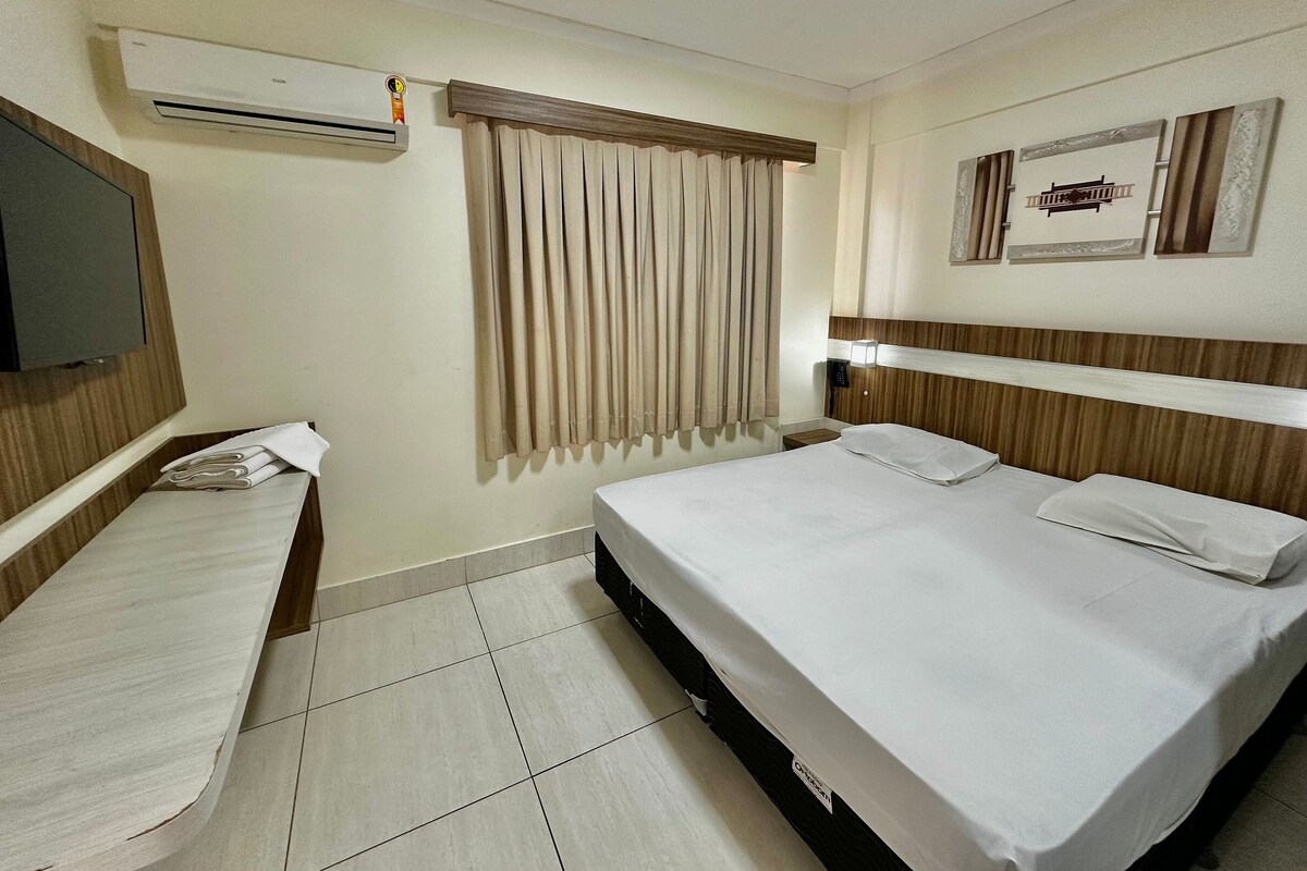 Habitación de hotel con cama, mesita de noche, televisor, ventana con cortinas y aire acondicionado instalado en la pared.
