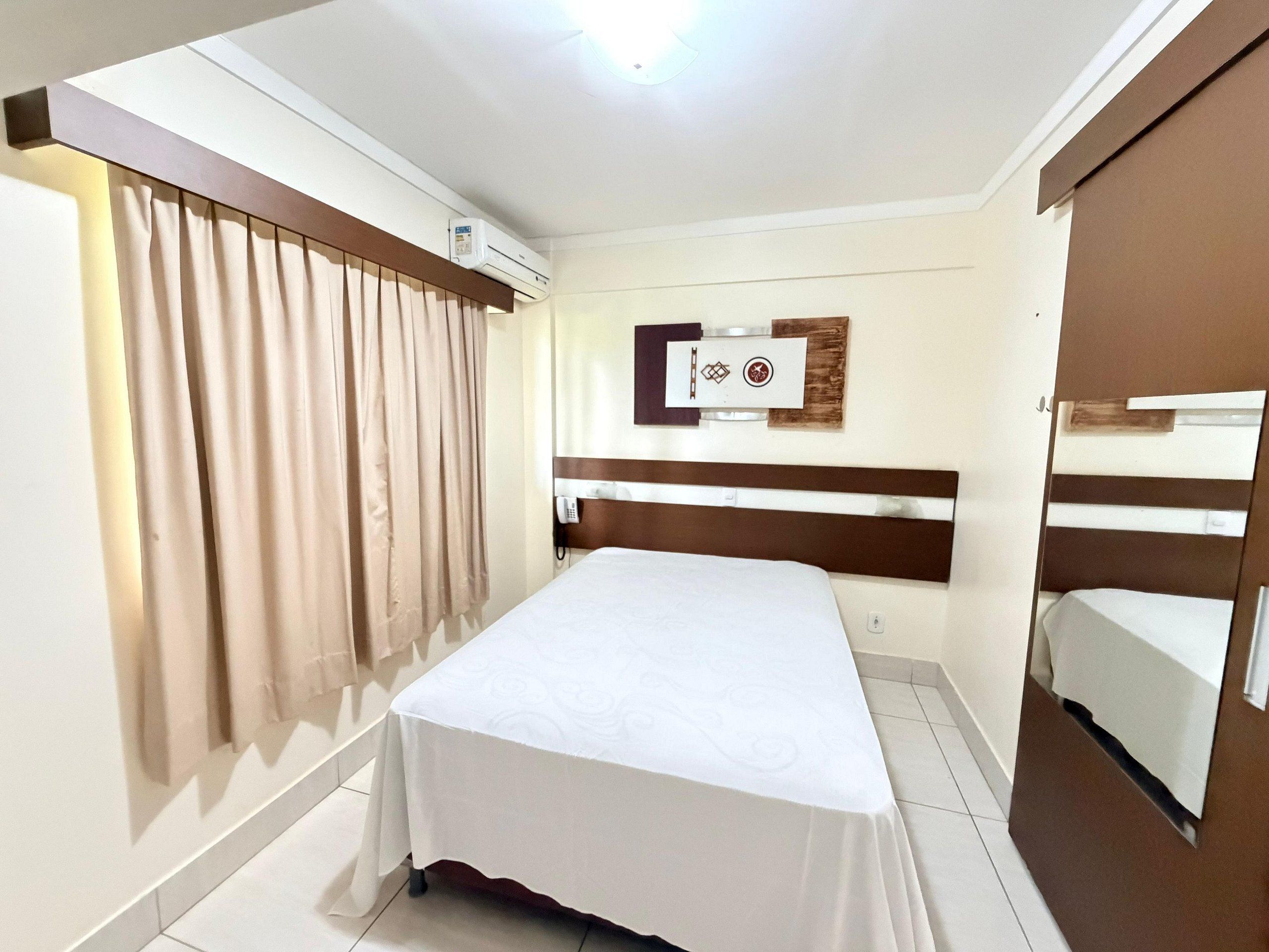 A hotel room with a bed, curtains, a wall-mounted air conditioner, a phone, and a wooden headboard.