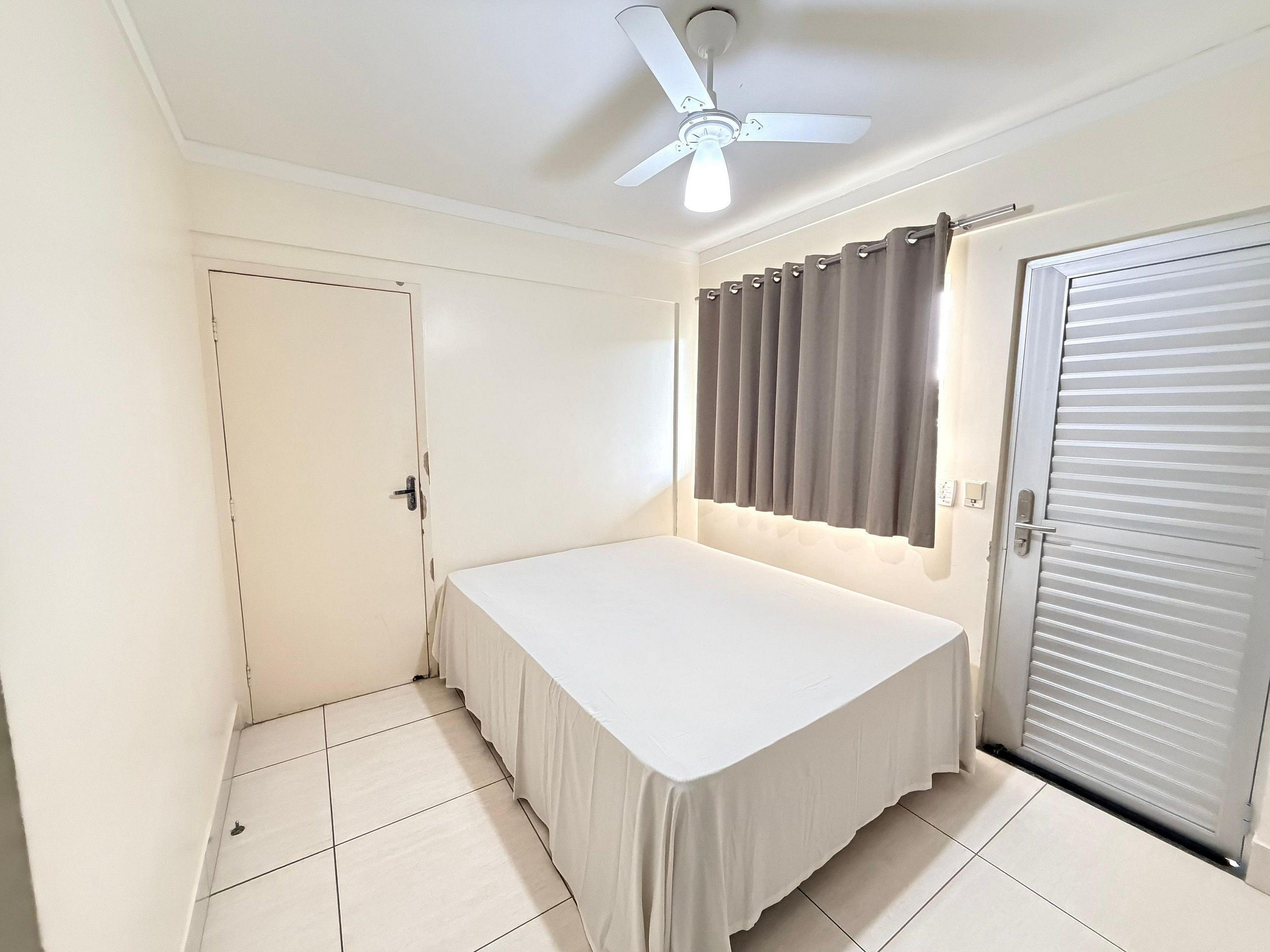 Une chambre à coucher avec un lit, un ventilateur de plafond, une fenêtre avec des rideaux et une porte.