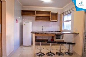 A compact kitchen with a refrigerator, microwave, and wooden cabinets. There are three bar stools and a window providing natural light. - Lacqua DiRoma V Via Caldas (Caldas Novas)