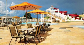 Un área junto a la piscina con sillones de descanso y mesas, piscina y estructura de tobogán acuático.