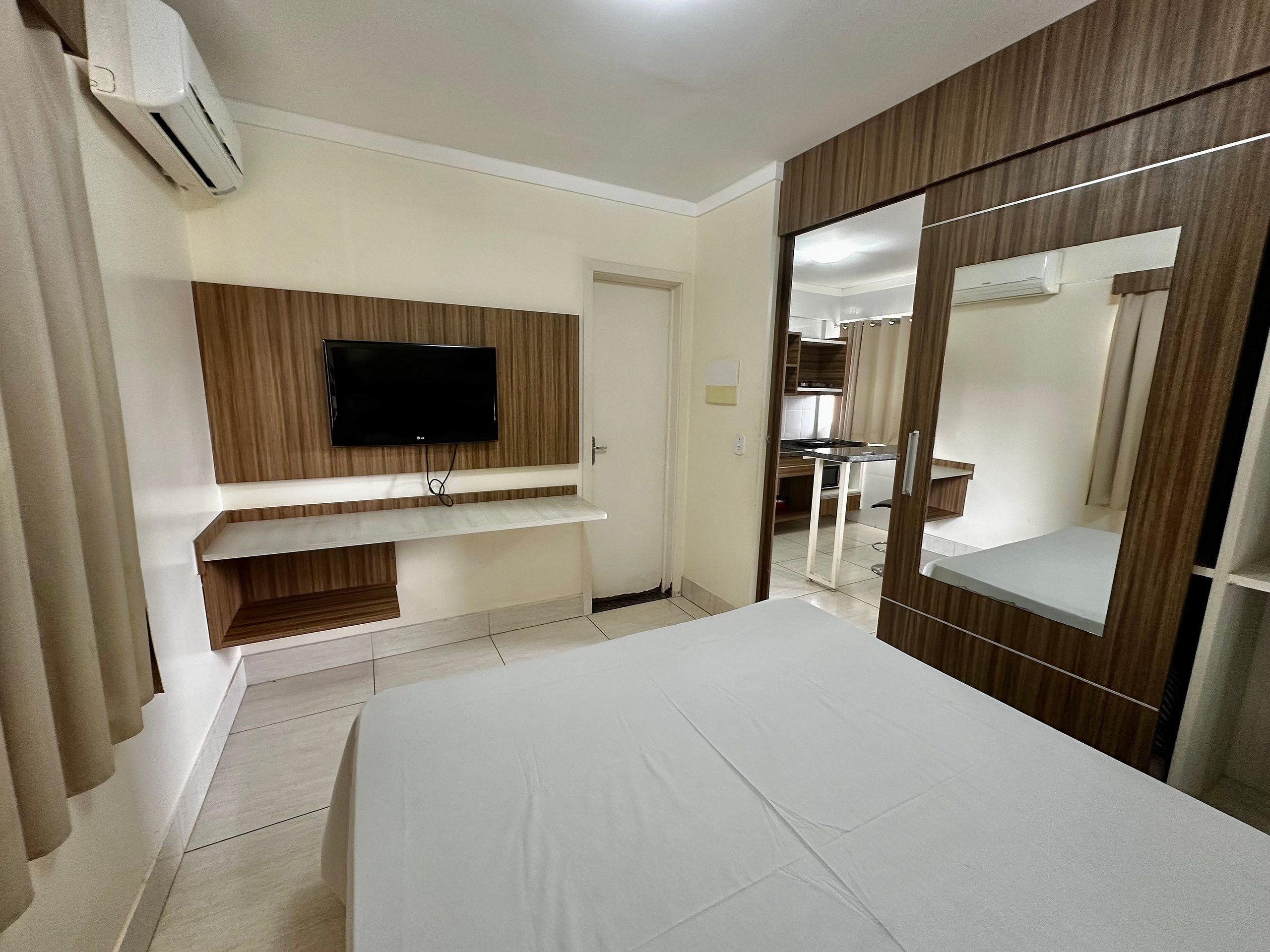 A hotel room with a bed, a TV mounted on a wooden panel, a wardrobe with a mirror, and an air conditioning unit.