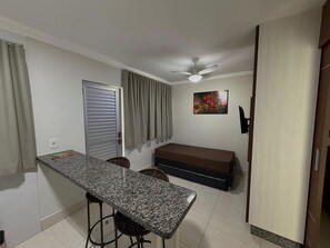 Una habitación compacta con kitchenette, ventilador de techo, cama y un cuadro en la pared.
