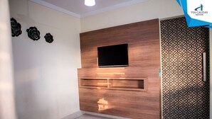 A modern living room with a flat-screen TV mounted on a wooden wall, a patterned decorative panel, and a tiled floor. - Lacqua DiRoma V Via Caldas (Caldas Novas)