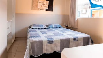 Quarto com cama, mesa de cabeceira, relógio na parede e janela com cortinas.