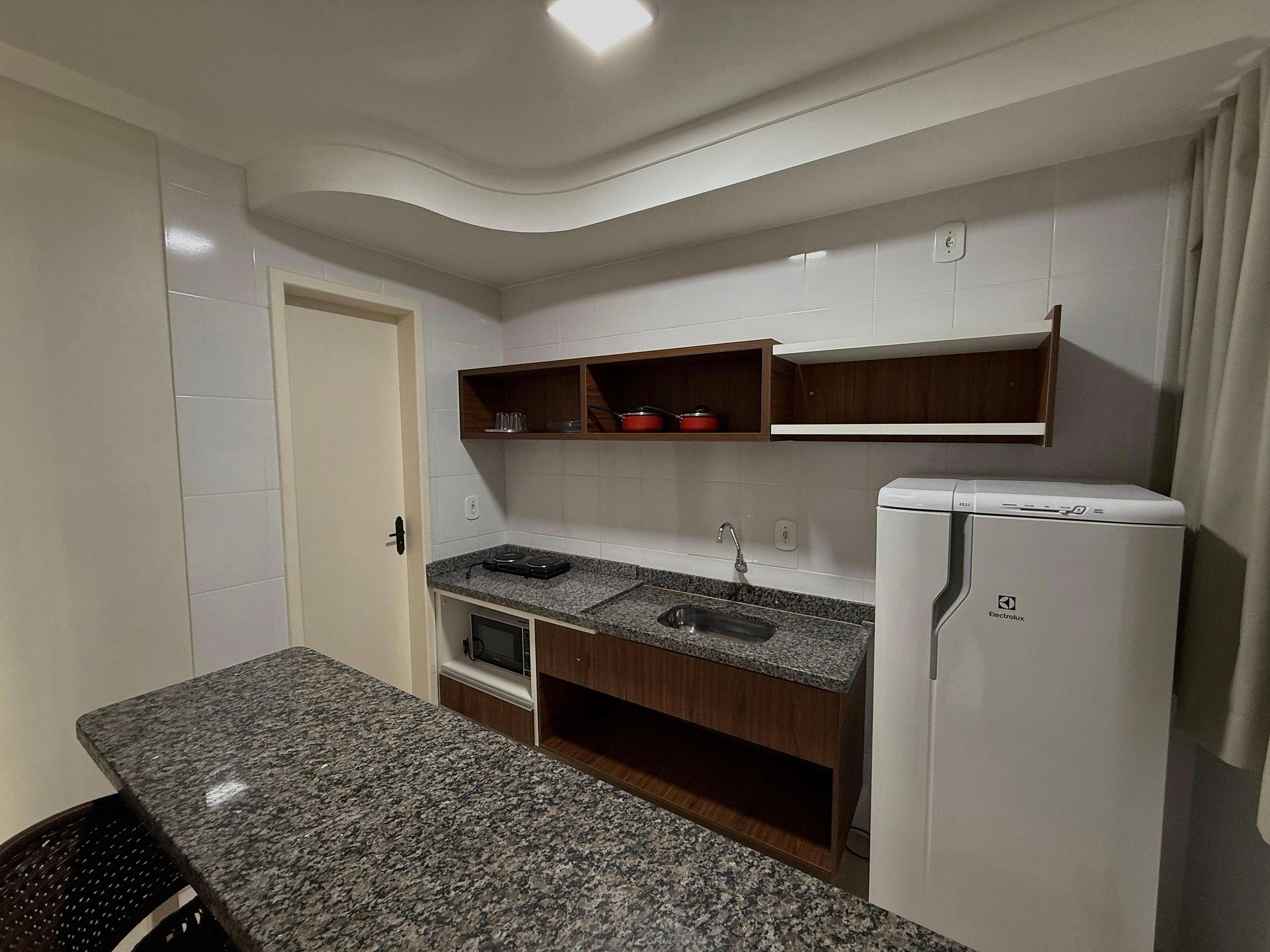 Una cocina compacta con encimera de granito, microondas integrado, refrigerador blanco y estantes de madera sobre el lavamanos.