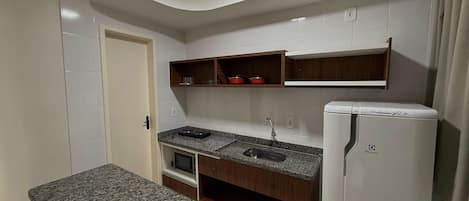 A compact kitchen with a granite countertop, a built-in microwave, a white refrigerator, and wooden shelves above the sink.
