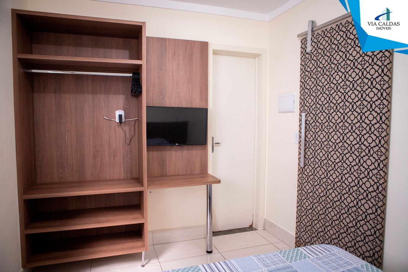 Una habitación con un armario de madera, un televisor en una estantería, una cortina estampada y suelo de baldosas.
