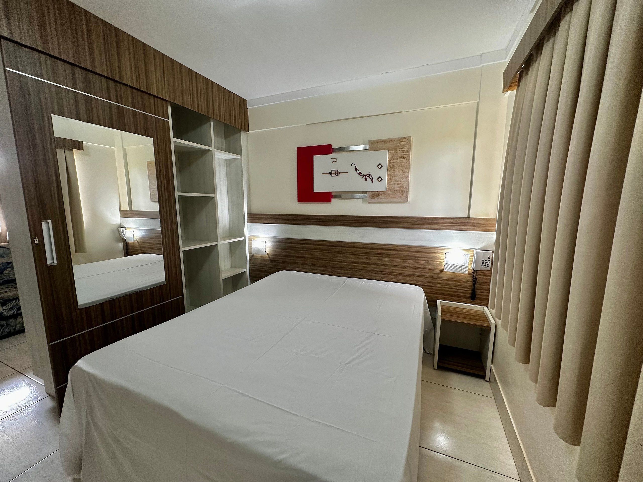 A hotel room with a bed, a wardrobe, a mirror, a clock, and a bedside table.