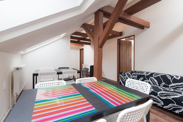 A cozy loft-style room with a sloped ceiling, a dining table with colorful striped tablecloths, white chairs, a black and white patterned sofa, and a wooden beam structure.