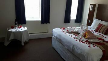A hotel room with a bed, a small table with a bottle and glasses, and a window with curtains.