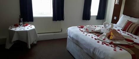Une chambre d’hôtel avec un lit, une petite table sur laquelle se trouvent une bouteille et des verres, et une fenêtre avec des rideaux.