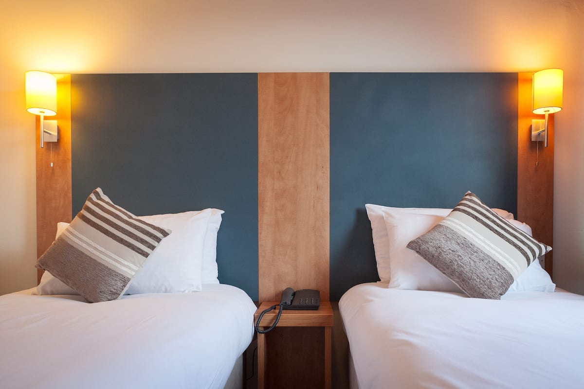Ein Hotelzimmer mit zwei Betten, jeweils mit einem Holzkopfteil, weißer Bettwäsche und gestreiften Kissen. An beiden Seiten des Zimmers befindet sich ein Nachttisch mit Telefon und einer an der Wand montierten Lampe.