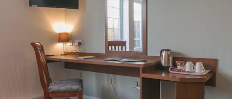 A hotel room with a desk, chair, TV, and a tray with cups and a kettle.